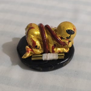 May include: A golden figurine of a figure lying down, with red accents and a black base. The figure is bound with a red cord. A white, cylindrical object is placed in front of the figure.