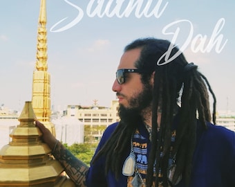 May include: A man with dreadlocks and a beard wears sunglasses and a blue shirt with a gold and blue patterned design. He is standing in front of a gold pagoda with a blue sky in the background. The text "Sadhu Dah" is written in white on the image.