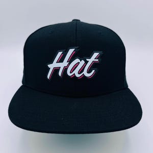 Puede incluir: Una gorra de b&eacute;isbol negra con un logotipo blanco "Hat" en un contorno rosa y blanco.