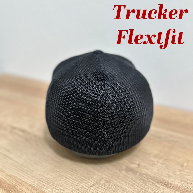 Puede incluir: Gorra de camionero negra con panel trasero de malla. La gorra tiene el texto "Flexfit" impreso en la parte delantera.