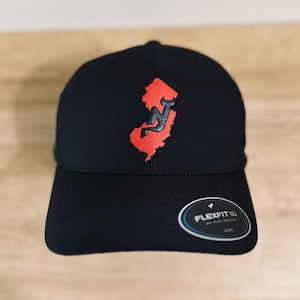 May include: Black baseball cap with a red outline of the state of New Jersey and a black "NJ" inside. The cap has a black Flexfit logo on the back.