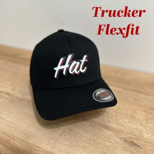 Puede incluir: Gorra de camionero negra con un logotipo "Hat" blanco en un contorno rojo. La gorra es de la marca Flexfit.