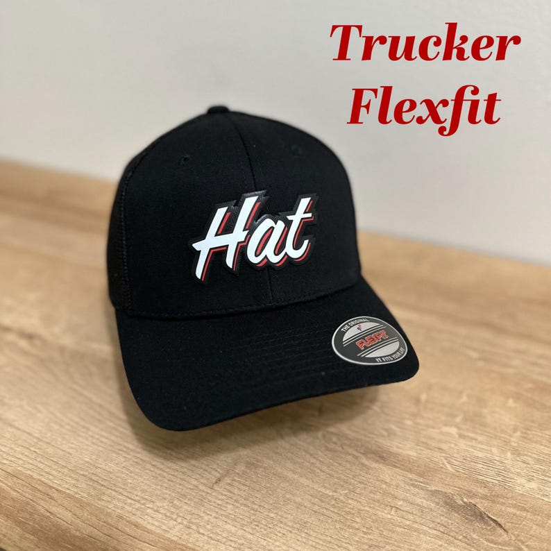 Puede incluir: Gorra de camionero negra con un logotipo "Hat" blanco en un contorno rojo. La gorra es de la marca Flexfit.