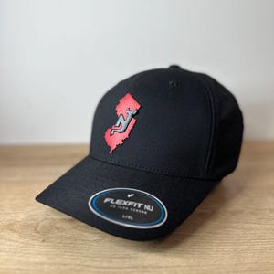 May include: Black baseball cap with a red and gray New Jersey outline logo. The cap has a Flexfit NU label on the underside of the brim.