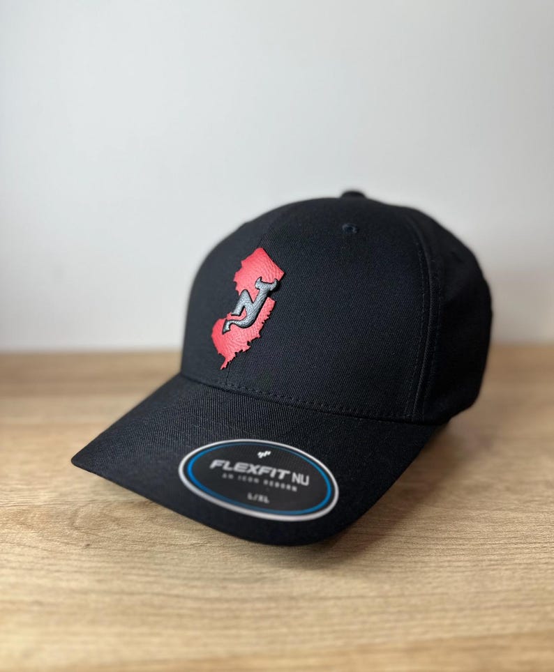 May include: Black baseball cap with a red and gray New Jersey outline logo. The cap has a Flexfit NU label on the underside of the brim.