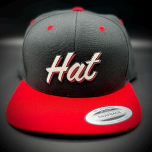 May include: A black and red snapback hat with the word "Hat" in white script. The hat has a red brim and red button on top. The hat has a "Snapback" tag.