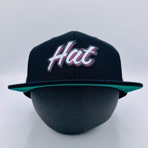 Puede incluir: Una gorra de b&eacute;isbol negra y verde con un logotipo blanco "Hat" en una fuente estilizada con acentos rojos.
