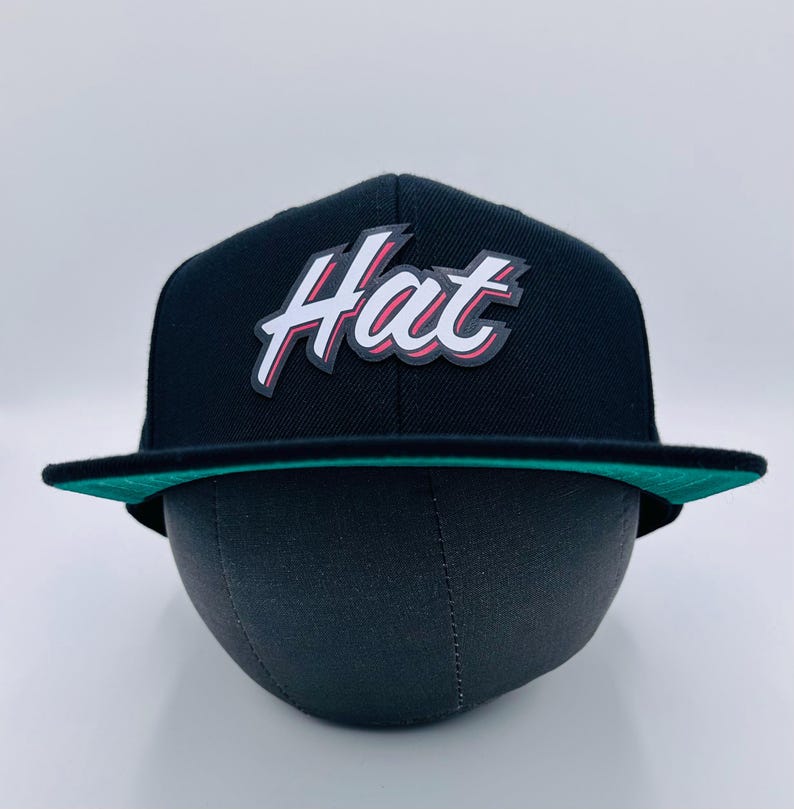 Puede incluir: Una gorra de b&eacute;isbol negra y verde con un logotipo blanco "Hat" en una fuente estilizada con acentos rojos.
