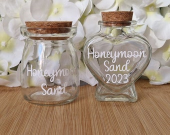 May include: Two clear glass jars with cork stoppers. One jar is round, the other is heart-shaped. Both jars have the words "Honeymoon Sand" in white script. The heart-shaped jar also has "2023" in white script. The jars are on a wooden surface.