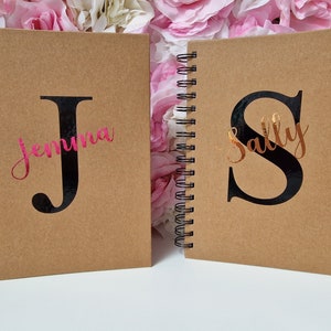 May include: Two spiral-bound notebooks with kraft paper covers. One notebook features a large black letter "J" with the name "Jemima" in pink script. The other has a black "S" with "Sally" in copper script.