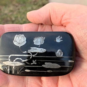 May include: A black lacquered box with a silver inlay design of water lilies and a bird. The box is rectangular and has a glossy finish.