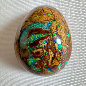 May include: A polished, oval-shaped gemstone with a brown, textured base and vibrant flashes of green, blue, and gold. The gemstone's surface is smooth and reflective, showcasing a unique pattern of colors.