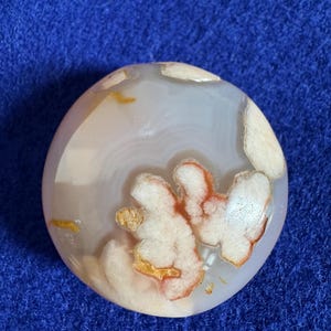 May include: A round, polished agate stone with a white, pink, and orange pattern. The stone has a smooth, polished surface and is a natural, organic material.