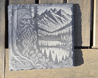 Puede incluir: Un posavasos rectangular de pizarra gris con un grabado de una escena de montaña y bosque. Una figura barbuda está en el lado izquierdo. El diseño es en tonos grises y blancos.