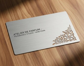 Puede incluir: Una tarjeta de visita rectangular de metal plateado con acabado cepillado. El texto ATELIER DE PARFUM - FRAGRANCE STUDIO - NEW YORK está impreso en negro. La esquina inferior derecha presenta un intrincado diseño floral cortado a láser.