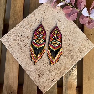 May include: A pair of long, beaded earrings with a geometric pattern in black, pink, orange, and green. The earrings have a fringe of black beads.