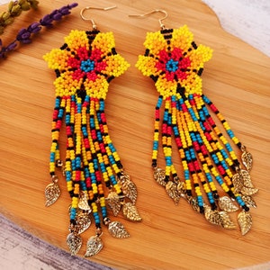 May include: A pair of long, beaded earrings with a star-shaped flower design in yellow, red, blue, and black. The earrings have gold leaf accents.
