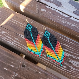 May include: A pair of long, beaded earrings with a black, turquoise, orange, and yellow colour scheme. The earrings are in the shape of a teardrop and have a geometric design at the top.