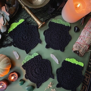 Pu&ograve; includere: Quattro sottobicchieri a forma di calderone in crochet nero con top verdi. I sottobicchieri sono disposti su una superficie verde con vari oggetti decorativi, tra cui candele, cristalli e un libro.
