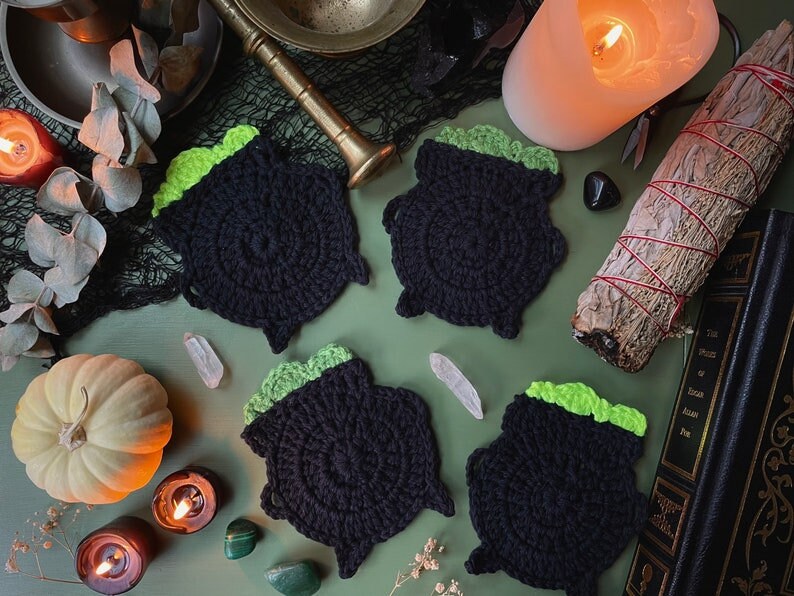 Pu&ograve; includere: Quattro sottobicchieri a forma di calderone in crochet nero con top verdi. I sottobicchieri sono disposti su una superficie verde con vari oggetti decorativi, tra cui candele, cristalli e un libro.