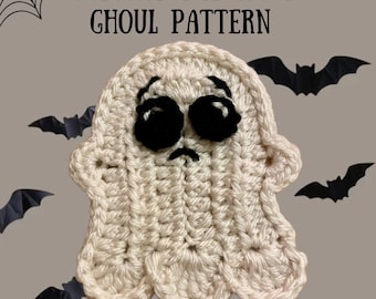 Può includere: Un fantasma all'uncinetto color crema con occhi neri e un'espressione triste, con la scritta "GREGORY THE GLUM GHOUL PATTERN". Il fantasma è circondato da ritagli di pipistrelli neri su sfondo grigio, evocando un tema di Halloween.