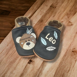 Könnte beinhalten: Ein Paar schwarze Lederschuhe für Babys mit goldenen Verzierungen. Ein Schuh ist mit einem gestickten Bären mit Federkopfschmuck verziert. Der andere Schuh ist mit dem Namen "Leo" bestickt.