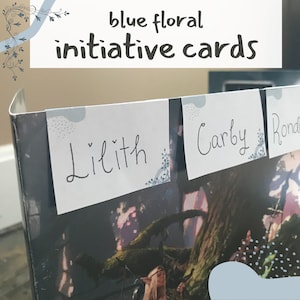 Könnte beinhalten: Nahaufnahme einer Schachtel mit drei weißen Karten, die oben angebracht sind. Die Karten tragen die Namen "Lilith", "Carby" und "Rond", die in schwarzer Tinte geschrieben sind. Die Schachtel hat ein blaues Blumenmuster und der Text "blue floral initiative cards" ist auf der Oberseite der Schachtel geschrieben.