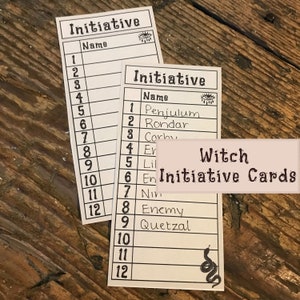 May include: Two white cards with black text and a black eye symbol. The cards are labeled "Initiative" and have numbered spaces for names. The top card has numbers 1-12. The bottom card has numbers 1-12 and has names written in the spaces, including "Penjulum", "Rondar", "Corh", "Er", "Li", "En", "NIV", "Enemy", and "Quetzal". A pink sticky note with the text "Witch Initiative Cards" is on the bottom card.