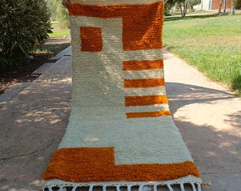 May include: A rectangular, hand-woven rug with a cream-colored background and geometric orange shapes. The rug features a textured, plush surface and a fringe along the bottom edge. The design includes a square and horizontal stripes.