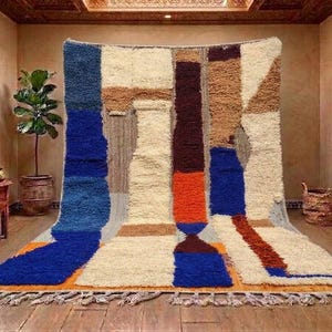 May include: A large, rectangular, hand-knotted wool rug featuring a bold, abstract geometric pattern. The design incorporates blocks of cream, deep blue, burnt orange, dark brown, and tan, finished with a fringed edge along the bottom.
