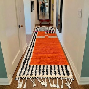 May include: A long rectangular runner rug featuring a vibrant orange central block, black and white striped borders, and a geometric pattern at one end. The rug is finished with white knotted fringe tassels along the bottom edge on a wooden floor.