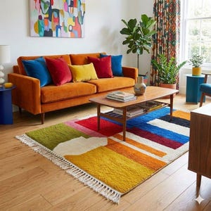 May include: A living room scene featuring an orange velvet sofa with colorful throw pillows. A low wooden coffee table sits on a vibrant, geometric rug with a fringed edge. The room is decorated with a colorful painting and potted plants.