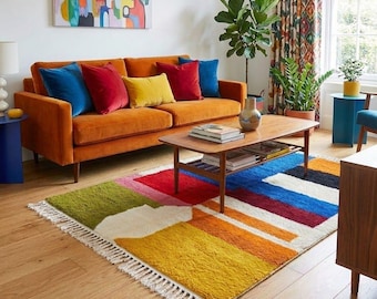 May include: A living room with an orange velvet sofa and colourful cushions. A low wooden coffee table rests on a vibrant, geometric rug with a fringed edge. An abstract painting hangs on the wall, enhancing the room's decor.