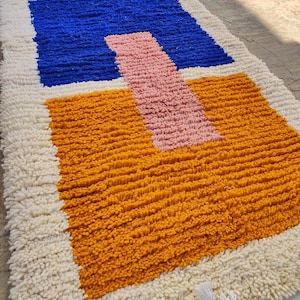 Könnte beinhalten: Rechteckiger Teppich mit geometrischem Design. Der Teppich hat ein blaues Quadrat, ein rosa Rechteck und ein orangefarbenes Quadrat, alles vor einem weißen Hintergrund. Der Teppich hat eine zottelige Textur und Fransen.