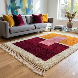 Può includere: Un tappeto rettangolare con base color crema caratterizzato da grandi blocchi geometrici in bordeaux intenso, arancione brillante e rosa tenue. Il tappeto presenta una consistenza spessa e folta, rifinita con lunghe frange annodate.