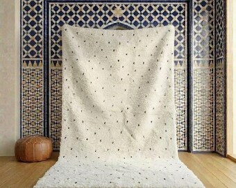 May include: A cream-colored, textured rug with small black dots and fringe along the bottom edge. The rug is displayed against a backdrop of patterned tiles in shades of blue, brown, and white, with a brown leather ottoman in the corner.