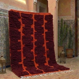Könnte beinhalten: Ein rechteckiger Teppich mit einem dicken, zotteligen Flor und einem sich wiederholenden geometrischen Muster aus dunkelburgunderroten Blöcken auf einem leuchtend orangefarbenen Hintergrund. Der Teppich hat eine weiße Fransenborte.