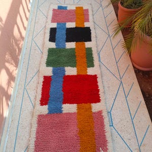 Op de afbeelding: Een rechthoekig tapijt met een levendig geometrisch patroon. Het ontwerp van het tapijt bevat blokken roze, oranje, blauw, zwart, groen en rood. Het ligt op een lichtgekleurd oppervlak met een blauw geometrisch patroon.