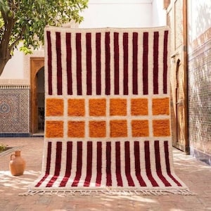 Peut inclure: Un tapis rectangulaire au fond crème orné d'un motif géométrique. Le design présente des rayures verticales rouge foncé sur les parties supérieure et inférieure, séparées par une bande horizontale centrale de dix carrés orange vif.