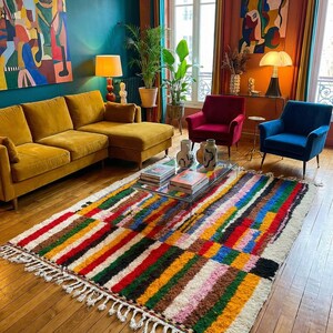 May include: A colorful, striped area rug with a white fringe. The rug features a variety of colors including red, green, yellow, and blue. The rug is placed in a living room with a yellow sofa, red and blue armchairs, and a glass coffee table.