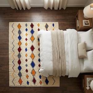 May include: A rectangular area rug with a cream-colored base featuring vertical rows of geometric diamond and zigzag patterns in blue, orange, and dark red. The rug is positioned next to a bed with white bedding and a beige textured throw blanket.