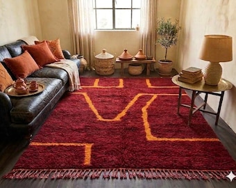 May include: A deep red rug featuring an orange geometric pattern. The rug has a thick pile and a fringed edge. The rug is in a living room setting with a leather sofa and decorative cushions. The rug measures approximately 200cm x 300cm.