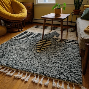 May include: A textured area rug with a geometric design. The rug features a dark gray and cream color palette with a pattern of black and cream shapes. The rug has a fringe of white tassels along the bottom edge.