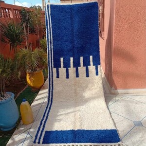 May include: A rectangular, blue and white rug with a geometric design. The rug features a large blue section at the top and bottom, with a white center. The design includes vertical blue stripes and a fringe at the bottom. The rug is made of a thick, textured material.