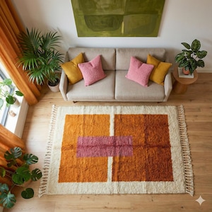 Könnte beinhalten: Draufsicht auf ein Wohnzimmer mit beigem Sofa, rosa und gelben Kissen und einem geometrischen Teppich. Der Teppich hat orange und rosa Blöcke mit cremefarbenem Rand und Fransen. Grüne Pflanzen und ein Gemälde ergänzen das Bild.