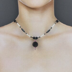 Peut inclure: Un collier de perles noires et blanches avec un pendentif en pierre de lave noire. Le pendentif est surmonté d'une petite perle rose.