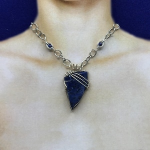 May include: A silver chain necklace with a triangular, dark blue stone pendant. The pendant is wrapped in silver wire. Small blue beads are interspersed along the chain.