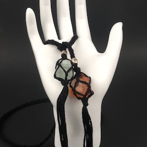 Könnte beinhalten: Eine schwarze Makramee-Halskette mit drei Kristallsteinen. Die Steine sind grün, orange und hellbraun. Die Halskette hat schwarze Quasten.