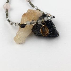 Puede incluir: Un collar de oro con un colgante de piedra preciosa marrón. El collar está hecho con pequeñas cuentas blancas, rosadas y negras. El colgante está rodeado por un marco de oro.