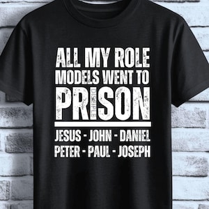 May include: A black short-sleeved t-shirt featuring white distressed text that reads ALL MY ROLE MODELS WENT TO PRISON followed by the names JESUS - JOHN - DANIEL - PETER - PAUL - JOSEPH. The brand name DISCIPLE Ink is printed in the top right corner.